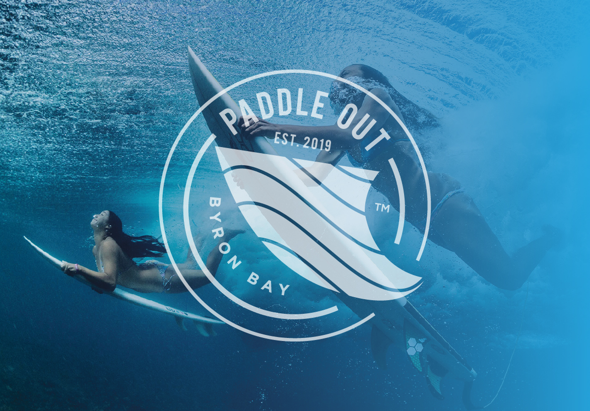 White outline version of the Paddle Out logo overlaid on an underwater shot of a surfer paddling, capturing the Byron Bay surf culture.